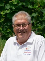 Ein lächelnder Mann mit Brille trägt ein weißes T-Shirt und sitzt vor üppigem Grün. Im Hintergrund sind Weinreben zu sehen.