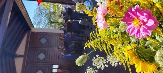 Eine bunte Blumenarrangement im Vordergrund mit verschiedenen Blumen. Im Hintergrund ist eine Gruppe von Menschen, die in einem geschlossenen Raum stehen.