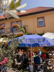Eine belebte Außenterrasse mit Tischen und Stühlen unter einem blauen Sonnenschirm des Lions Clubs. Im Hintergrund sind ein gelbes Gebäude und grünes Laub zu sehen.