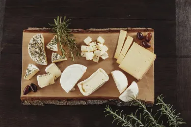 Eine Auswahl an verschiedenen Käsesorten auf einem Holzbrett. Neben dem Käse befinden sich einige Oliven und frische Kräuter.