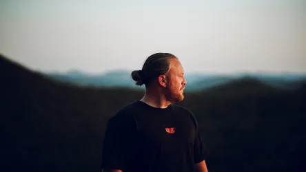 Ein Mann mit einem Pferdeschwanz steht auf einem Hügel. Im Hintergrund sind sanfte Berge und ein klarer Himmel zu sehen.