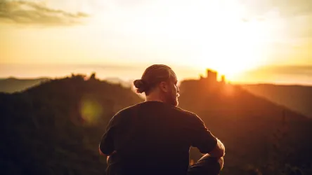 Ein Mann sitzt auf einem Berg und blickt auf einen schönen Sonnenuntergang. Die Landschaft ist grün und hügelig, mit einem alten Gebäude im Hintergrund.