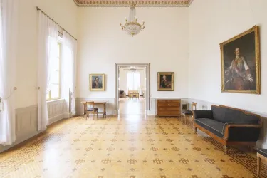 Ein eleganter Raum mit Holzboden und hohen Decken. An den Wänden hängen Porträts, und in der Mitte steht ein Sofa sowie ein Tisch.