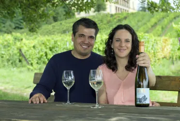 Ein Paar sitzt an einem Tisch im Freien und hält eine Flasche Wein. Im Hintergrund sind Weinreben zu sehen.