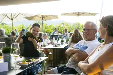 Eine fröhliche Gruppe von Menschen in einem Außenbereich. Eine Person schenkt Wein ein und alle genießen das Essen und die Atmosphäre.