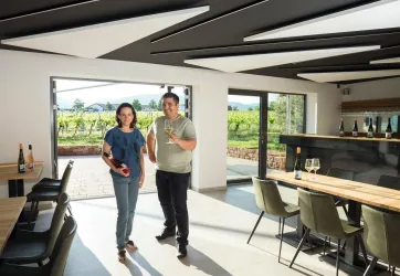 Ein Weinliebhaber zeigt stolz seine Weinsammlung in einem modernen Weingut. Im Hintergrund sind Weinberge und eine helle, einladende Atmosphäre zu sehen.