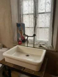 Ein Waschbecken aus Keramik steht auf einem Holzuntergrund. Im Hintergrund sind ein Fenster mit Vorhängen und ein dekoratives Bild zu sehen.