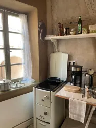 Eine gemütliche Küche mit einem alten Herd, einer Kaffeemaschine und einem Fenster, das viel Licht hereinscheinen lässt. Auf dem Regal sind verschiedene Küchenutensilien und Ölflaschen sichtbar.