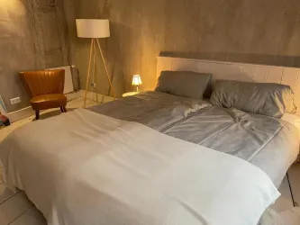 Ein gemütliches Schlafzimmer mit einem großen Bett und grauer Bettwäsche. Im Raum stehen eine Stehlampe und ein bequemer Sessel aus braunem Leder.