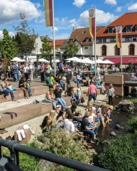 Ein belebter Platz mit vielen Menschen, die sich im Freien entspannen. Im Hintergrund sind Cafés und grüne Bäume zu sehen.