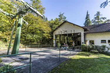 Eine Seilbahnstation im Wald mit einem großen Gebäude und grüner Natur umgeben. Im Vordergrund warten einige Besucher auf die Seilbahn.