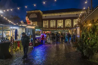 Ein festlicher Weihnachtsmarkt in der Dämmerung, beleuchtet von Lichtern. Besucher schlendern zwischen den Ständen und genießen die Atmosphäre.