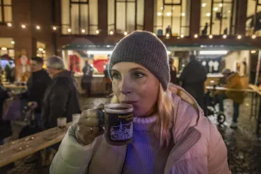 Eine Frau trinkt aus einer Tasse, während sie auf einem festlichen Markt steht. Im Hintergrund sind Lichter und Menschen zu sehen.