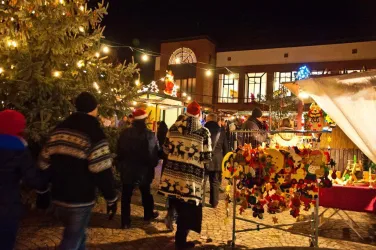 Ein festlicher Weihnachtsmarkt mit Lichtern und Ständen. Menschen in winterlicher Kleidung genießen die Atmosphäre und stöbern nach Geschenken.