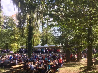 Eine belebte Outdoor-Veranstaltung mit vielen Menschen, die an Tischen sitzen. Im Hintergrund sind Zelte und Bäume zu sehen.