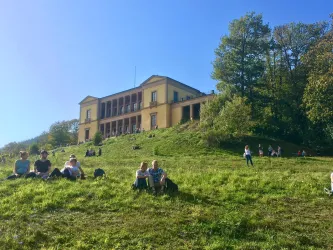 Ein historisches Gebäude steht auf einer grünen Wiese. Menschen sitzen im Gras und genießen die Sonne.