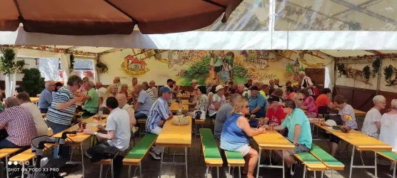 Eine große Gruppe von Menschen sitzt an langen Tischen unter einem Zelt. Die Atmosphäre ist lebhaft und einladend.