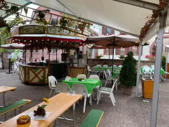 Ein gemütlicher Innenhof mit Tischen und Stühlen unter Schirmen. Im Hintergrund steht ein kleiner Verkaufsstand mit festlicher Beleuchtung.