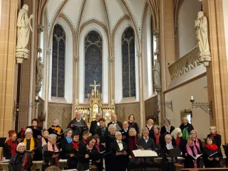 Ein Chor steht in einer Kirche und singt. Im Hintergrund sind bunte Fenster und religiöse Statuen zu sehen.