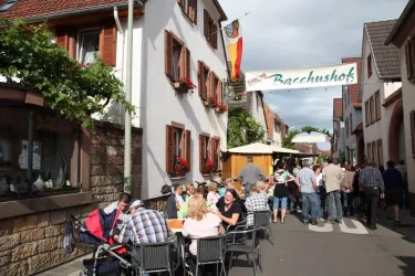 Eine belebte Straße mit Menschen, die an Tischen sitzen und essen. Im Hintergrund sieht man traditionelle Häuser und eine Fahne.