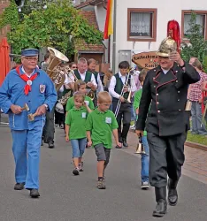 Eine festliche Parade mit Menschen in traditionellen Kleidern und Kindern in grünen T-Shirts. Musikalische Gruppen und eine fröhliche Atmosphäre prägen die Szene.