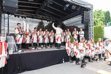 Eine Gruppe von Musikern in historischen Uniformen steht auf einer Bühne. Sie präsentieren ein musikalisches Stück vor einem Publikum.