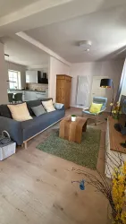 Ein modernes Wohnzimmer mit einem grauen Sofa und einem Holztisch. Der Raum hat Holzfußboden und große Fenster, die für viel Licht sorgen.