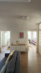 Ein Wohnzimmer mit einem grauen Sofa und einem weißen Sideboard. Im Hintergrund sind zwei Schlafzimmer mit Fenstern und hellem Licht sichtbar.