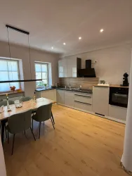 Eine moderne Küche mit einem Essbereich. Die Wände sind hell, und es gibt große Fenster für viel Tageslicht.