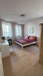 Ein gemütliches Schlafzimmer mit einem Doppelbett und roten Bettlaken. Das Zimmer ist hell und freundlich, mit großen Fenstern und moderne Möbeln.