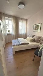 Ein helles Schlafzimmer mit einem Doppelbett und sanfter Bettwäsche. Große Fenster lassen viel Tageslicht herein und sorgen für eine angenehme Atmosphäre.