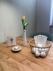 Ein schöner Tisch mit einem weißen Vasen und einer gelben Tulpe. Daneben sind Karten und ein Korb mit Eiern platziert.