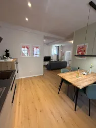 Eine moderne Küche mit Holzfußboden und einem Essbereich. Im Hintergrund ist ein gemütliches Wohnzimmer mit einem Sofa und Fernseher zu sehen.