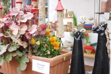 Ein bunter Blumentopf mit verschiedenen Pflanzen und einer kleinen roten Türmchen-Deko. Daneben stehen zwei schwarze Figuren, die Katzen darstellen.