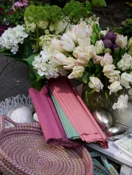 Ein schöner Blumenstrauß mit weißen und bunten Tulpen liegt neben bunten Tuchrollen und einem geflochtenen Korb. Die Szene vermittelt eine fröhliche und farbenfrohe Atmosphäre.