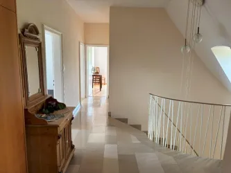 Ein heller Flur mit Treppe und eleganten Leuchten. Auf der linken Seite steht ein Holzschrank und im Hintergrund ist ein Tisch sichtbar.