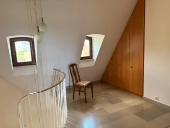 Ein gemütlicher Raum mit einer Holzstuhlle und schrägen Fenstern. Die Wände sind hell und der Boden ist gefliest.