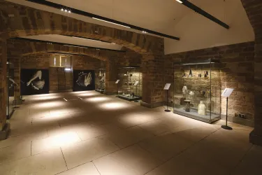 Ein Innenraum eines Museums mit Steinwänden und mehreren Vitrinen. In den Vitrinen sind verschiedene Ausstellungsstücke zu sehen.