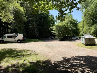 Ein Campingplatz im Wald mit mehreren Stellplätzen für Wohnmobile. Die Umgebung ist grün und sonnig, umgeben von Bäumen.