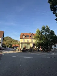 Ein charmantes Gebäude an einer Straßenecke mit roten Dächern und viel grünem Laub. Im Hintergrund sind sanfte Hügel und ein blauer Himmel sichtbar.