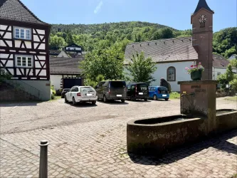 Ein ruhiger Dorfplatz mit historischen Fachwerkhäusern und einer kleinen Kirche. Im Vordergrund sind einige Autos geparkt und Bäume umgeben den Platz.