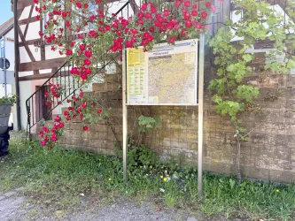 Ein Wegweiser mit einer Karte steht an einer Wand, umgeben von bunten Blumen. Im Hintergrund sieht man eine Treppe und einen Teil eines Hauses.