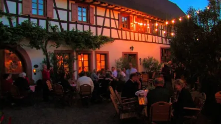 Eine lebhafte Terrasse mit vielen Gästen bei Abendlicht. Der Fachwerkbau im Hintergrund ist mit Lichtern geschmückt.