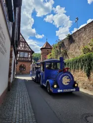 Ein blauer Touristenzug fährt durch eine malerische, enge Gasse. Im Hintergrund sind traditionelle Fachwerkhäuser und ein kleiner Turm sichtbar.