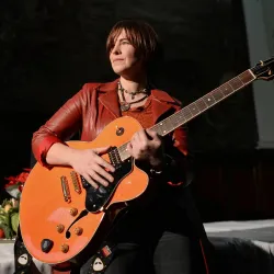 Eine Frau mit einer orangen Gitarre steht auf der Bühne. Sie trägt eine rote Jacke und hat eine selbstbewusste Ausstrahlung.