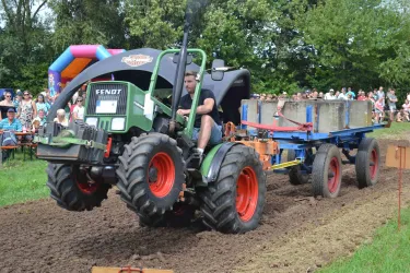 Ein Traktor zieht einen Anhänger mit Betonblöcken auf einem Feld. Im Hintergrund sind Zuschauer zu sehen, die das Spektakel beobachten.
