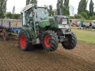 Ein grüner Traktor zieht eine landwirtschaftliche Maschine über einen bearbeiteten Acker. Im Hintergrund sind einige Bäume und Zuschauer zu sehen.