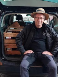 Ein Mann sitzt am Heck seines Autos, das mit Holzkisten gefüllt ist. Er trägt einen Strohhut und lächelt.