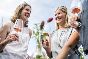 Zwei Frauen lächeln und genießen Roséwein in der Hand. Im Hintergrund sind Blumen zu sehen, was eine festliche Atmosphäre schafft.