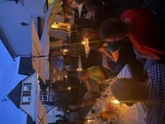 Eine gemütliche Abendessen-Atmosphäre im Freien, beleuchtet von Kerzen und Lampen. Die Tische sind mit weißen Tischtüchern gedeckt und umgeben von Gästen, die entspannt plaudern.